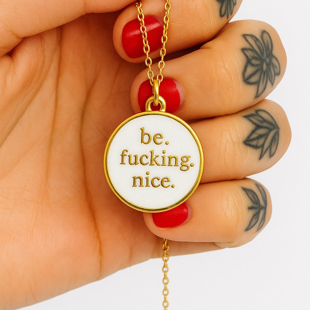Gold necklace with a pendant that says 'be. fucking. nice.' held by a hand.
