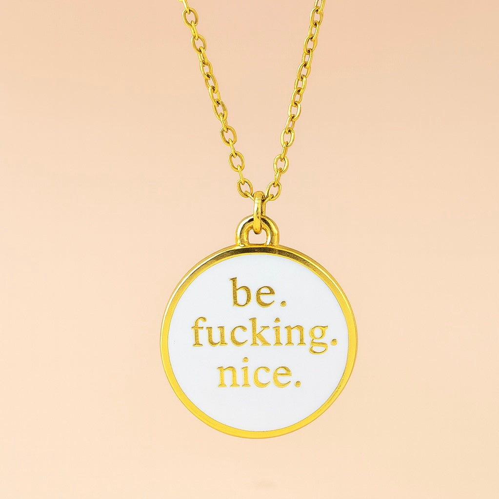 Gold necklace with a pendant that says 'be. fucking. nice.' on a beige background