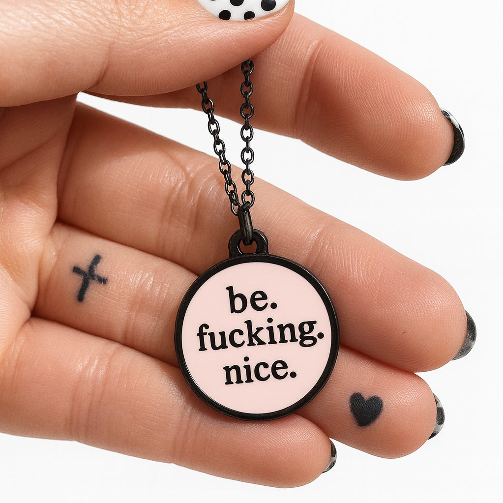 Black and pink Necklace with a pendant that says 'be. fucking. nice.' held by a hand.