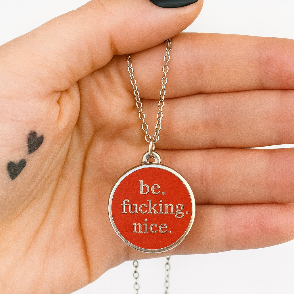 Necklace with a red pendant that says 'be. fucking. nice.' held in a hand.
